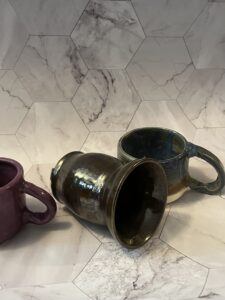 The image shows the same three clay pieces against the background, but this time the mugs are turned to show the handles are upside down and the vase is laying on its side because it can't stand without being leaned up against something.